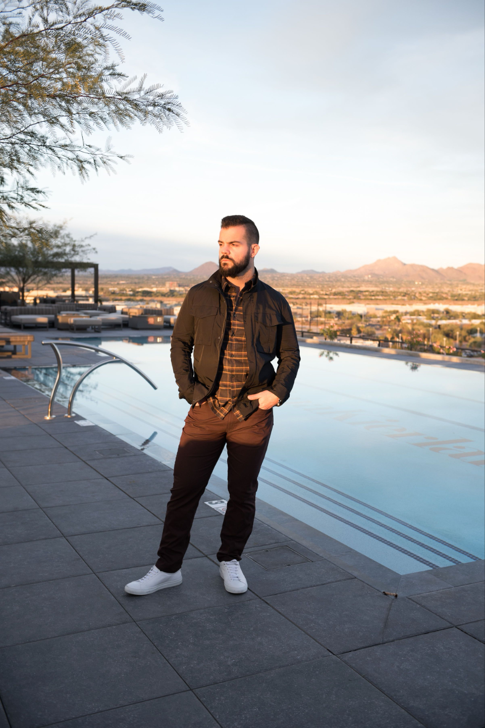 A fashionable man stands in front of pool overlooking city background. His hands are in his pockets. He wears a plaid shirt, brown pants, and white tennis shoes.